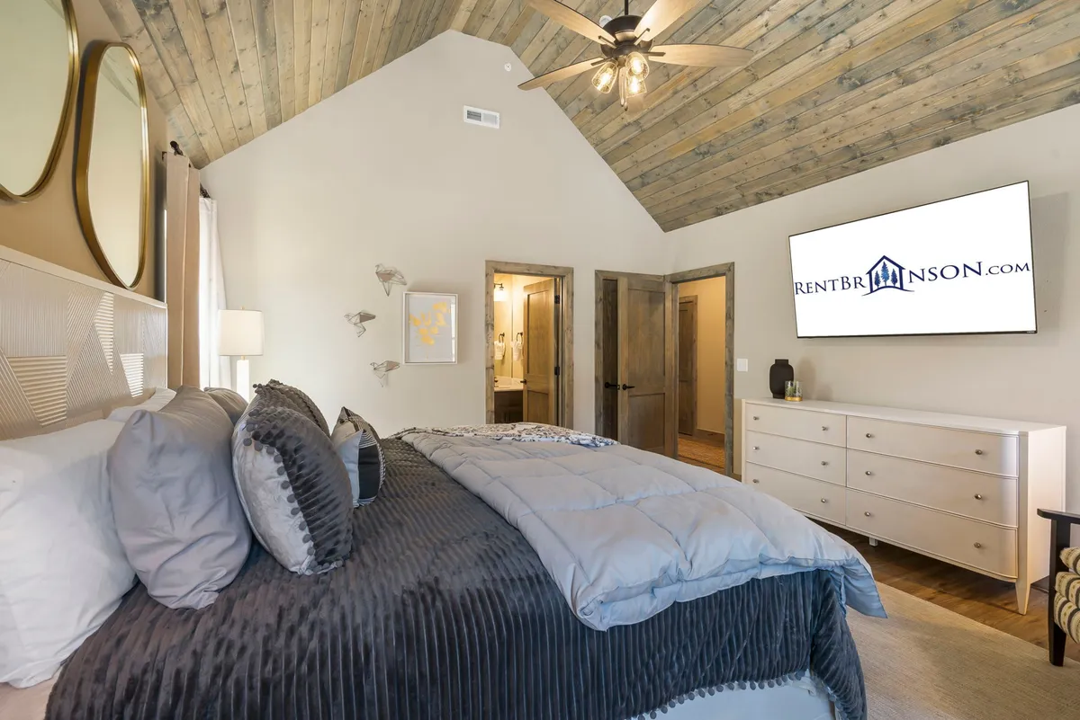 A king-sized bed with plush bedding sits against a wooden accent wall, complemented by decorative pillows. A large screen TV and dressers add comfort and function, while the wooden ceiling and soft lighting create a warm, serene ambiance.