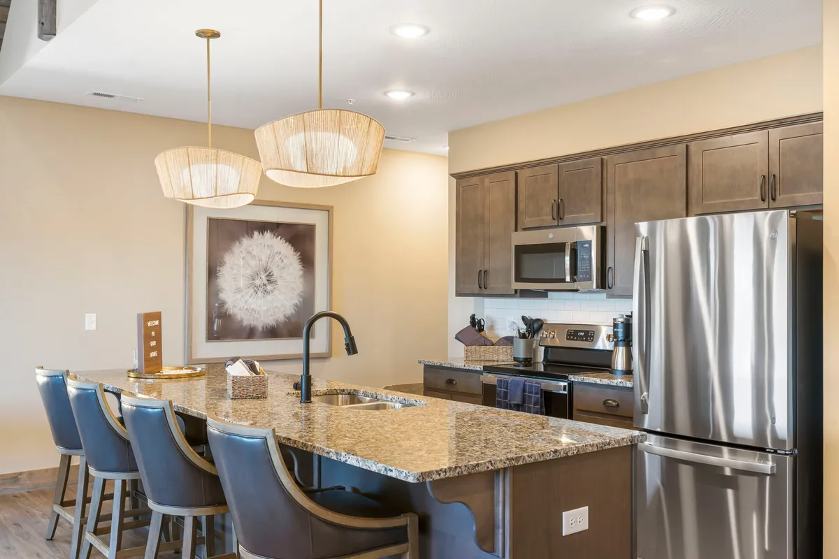 A bright, welcoming kitchen scene with a granite countertop, pendant lights, bar stools, and stainless steel appliances. The arrangement is ideal for casual dining and group cooking, creating an inviting ambiance for gathering and celebration.