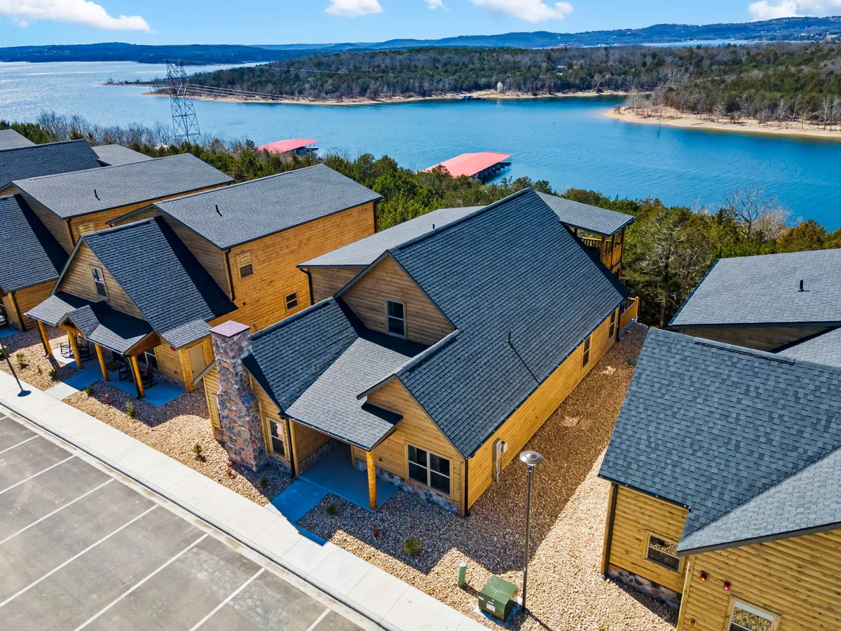 A collection of rustic wooden cabins nestles along the gentle curve of Table Rock Lake, with a wide expanse of water visible beyond and lush landscaping framing the scene. The foreground invites viewers to imagine outdoor gatherings, boating, and lakeside relaxation, all set against the backdrop of a stone chimney and open green spaces.