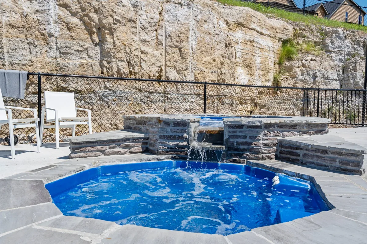 An exterior scene featuring a hot tub with a waterfall feature, surrounded by stone seating and landscaping. The area offers sunset views and an intimate, spa-like space for relaxation.