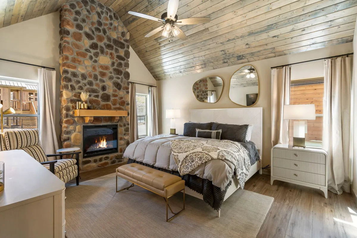 A tranquil master bedroom with a stone fireplace, large windows streaming natural light, a cozy bed with decorative bedding, and a striped chair. The space is ideal for relaxing, reading, or enjoying a quiet moment by the fire.