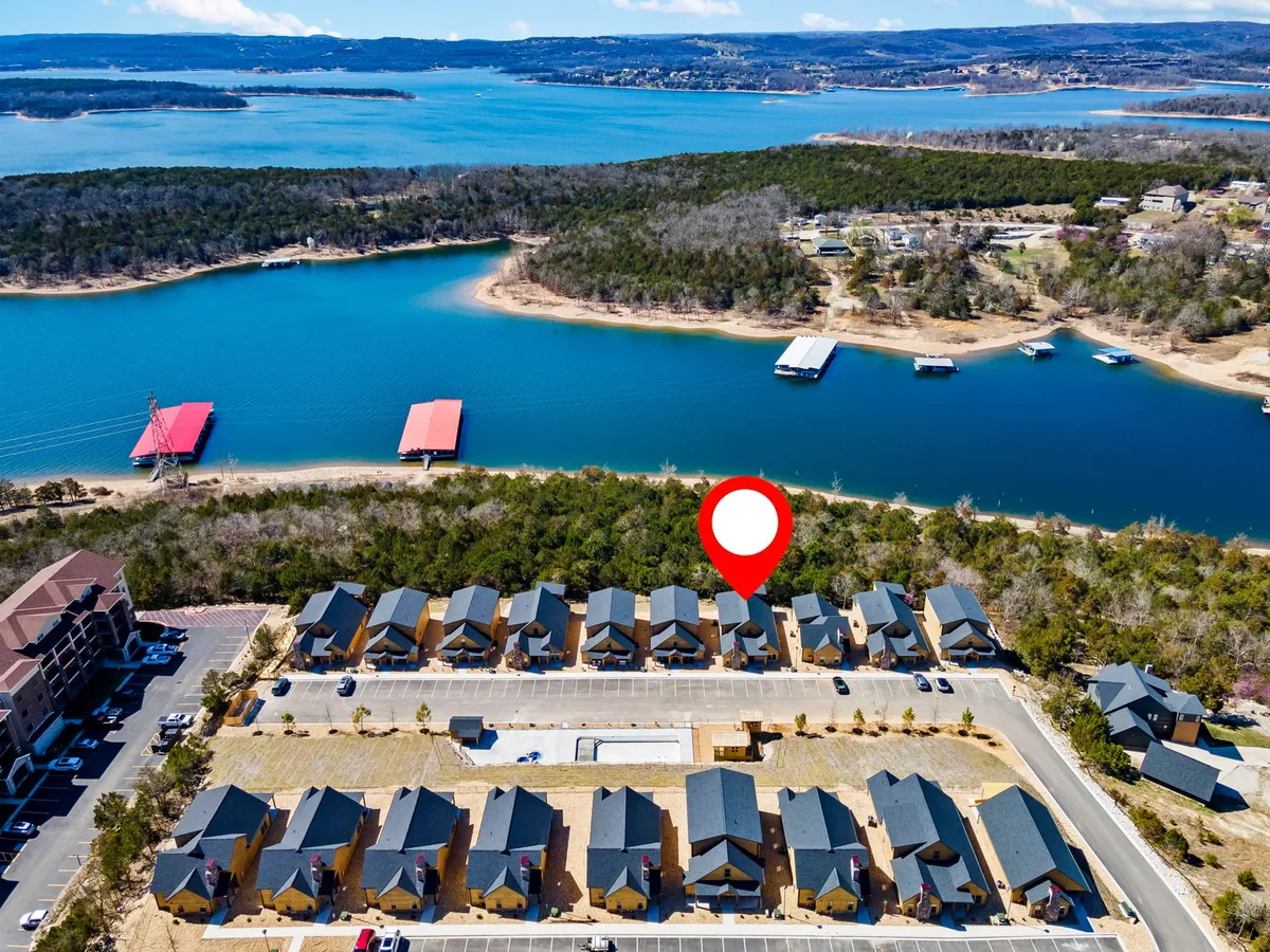 An exterior view of the lake with a row of homes nestled along the shoreline, surrounded by lush greenery and boathouses. The foreground features the tranquil waters of the lake and nearby parking for easy access.