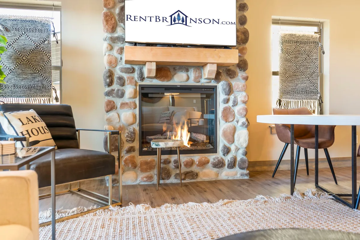 A warm, open living space with a comfortable seating area centered around a stone fireplace. The dining table is set nearby, all within a single, cohesive space, highlighting the open-plan layout. The natural light and cozy furnishings enhance the welcoming mood.