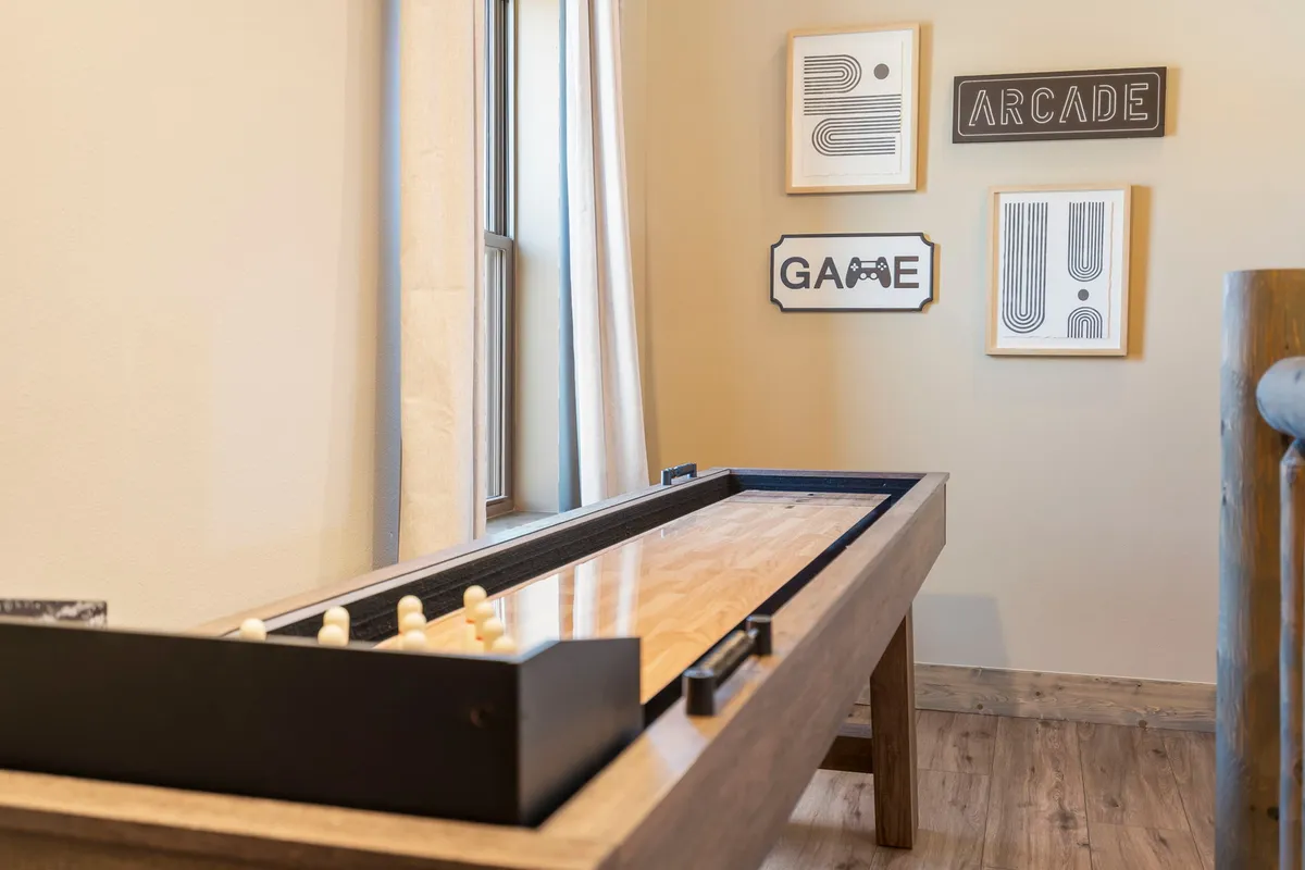 A well-lit loft area with a sliding game table as the focal point, framed wall art for character, and a large window with curtains. The scene is styled for socializing and relaxation, capturing a moment where family or friends can gather for games and conversation.
