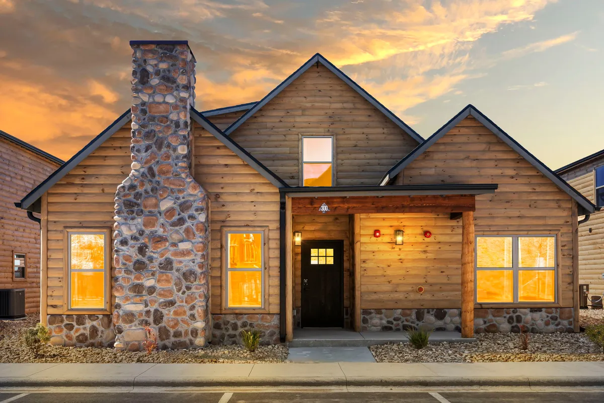 A welcoming exterior shot of a wooden cabin with a stone chimney, spacious front porch, and large windows. The scene evokes warmth and hospitality, ideal for gatherings or peaceful moments outdoors.