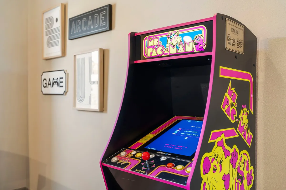 A vibrant, nostalgic corner featuring a Ms. Pac-Man arcade machine, framed wall art, and arcade-themed signage, styled for playful entertainment. The setup invites guests into a space ideal for friendly gaming tournaments and shared laughter.