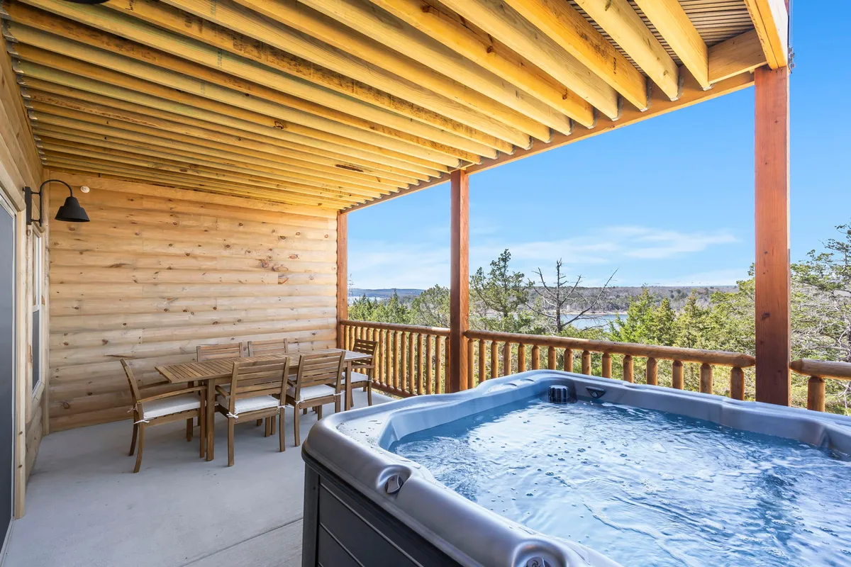 This exterior shot captures a covered patio area featuring a private hot tub, a dining table perfect for outdoor meals, a sturdy wooden railing, and a scenic view of the surrounding landscape. The hot tub is centrally placed for relaxation, while the open-air setup invites lakeside breezes.