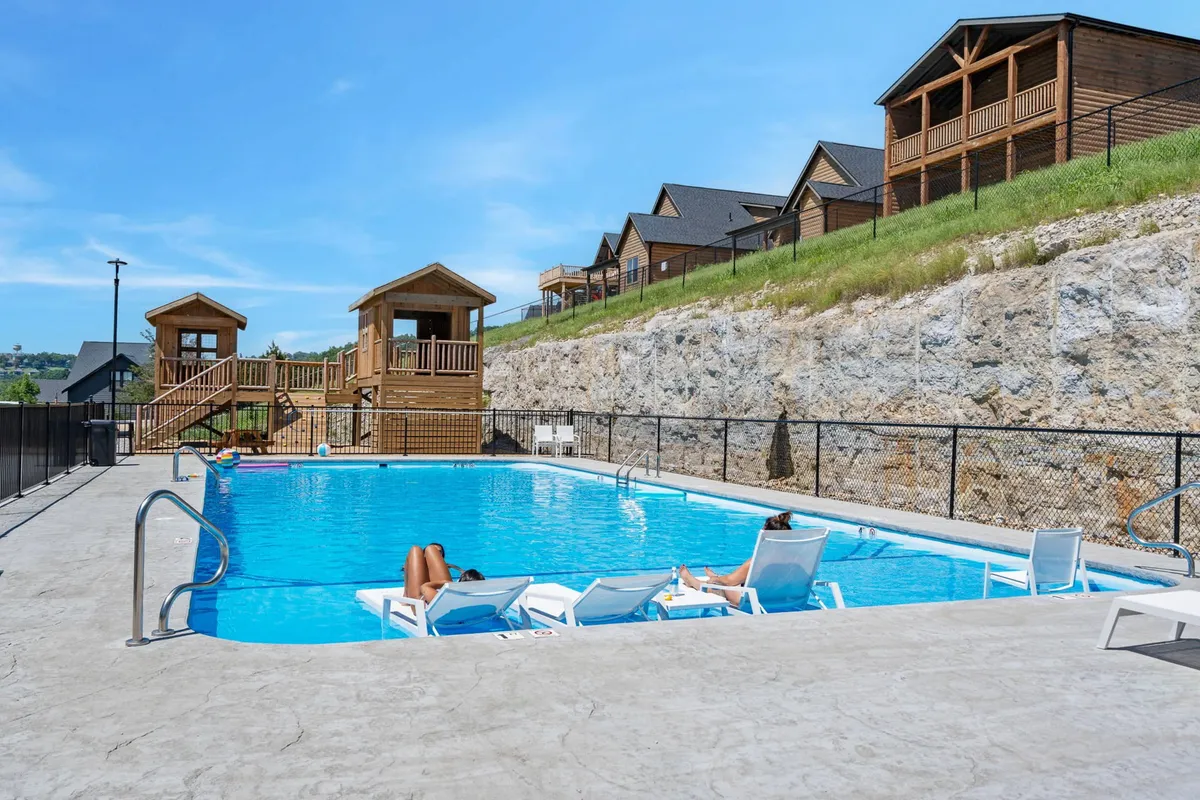 A vibrant, sunlit outdoor pool is surrounded by poolside loungers and a colorful playground. The scene is lively, with plenty of space for families to swim, sunbathe, and children to play, all set within a well-maintained resort environment.