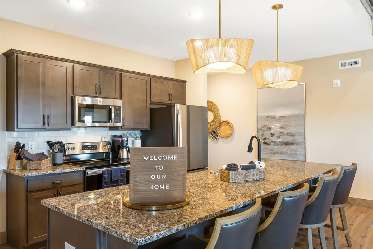 This image features a granite kitchen island surrounded by bar stools, pendant lighting overhead, and a signboard reading 'WELCOME TO OUR HOME.' The setting is warm and inviting, capturing the spirit of gathering and sharing meals together. The kitchen is well-lit and open, hinting at a seamless connection to other living areas.