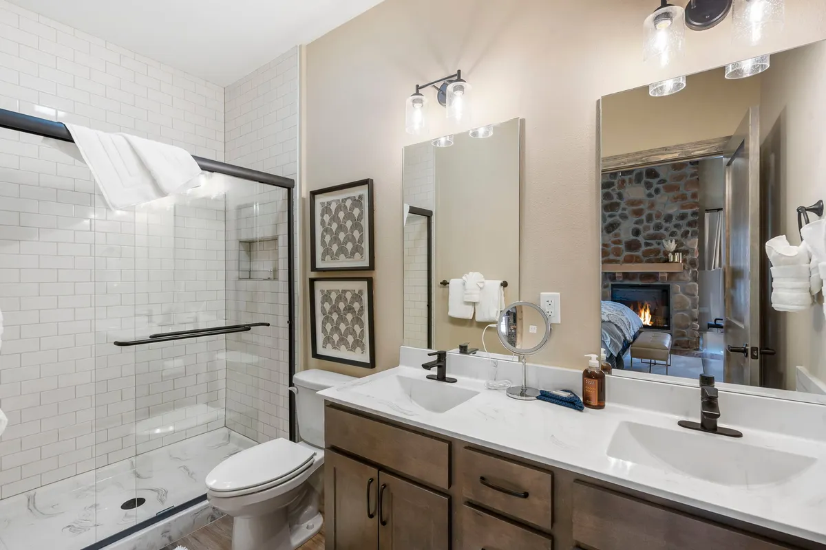 A modern, spacious bathroom featuring a glass-enclosed walk-in shower, double sink vanity with ample counter space, a mirrored wall, and a glimpse of a stone fireplace in the background. The space is bright and serene.